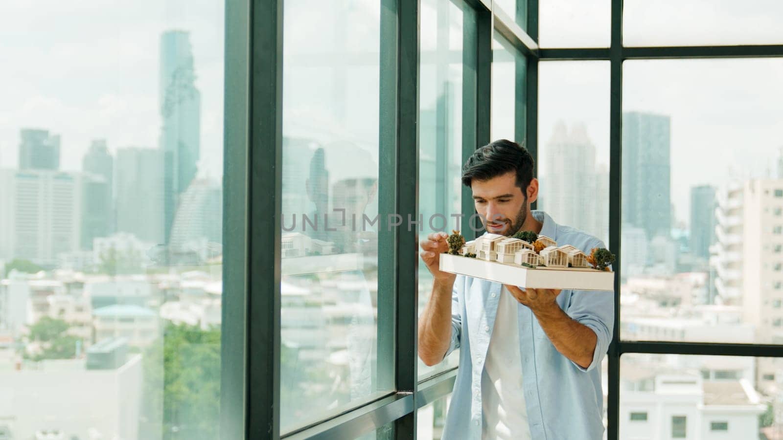 Businessman in casual outfit holding house model while checking house construction. Architect engineer inspect building model while standing near window with skyscraper. Engineering. Tracery