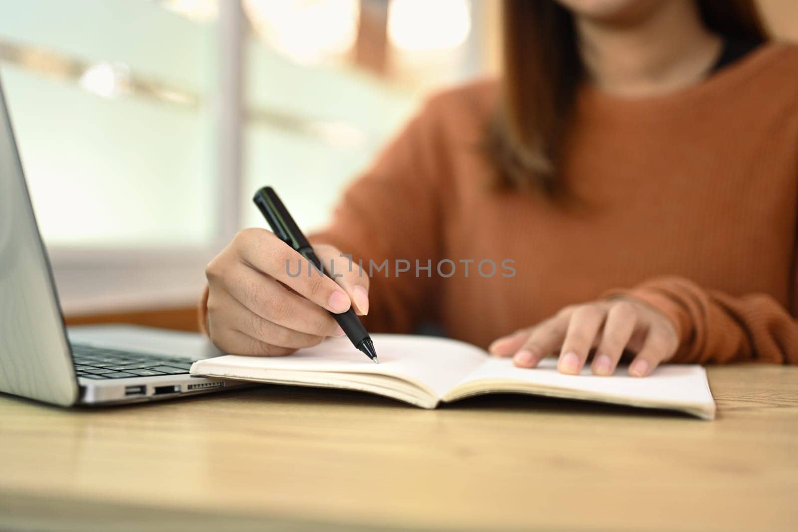 Cropped shot of young woman making important notes and using laptop on wooden desk.