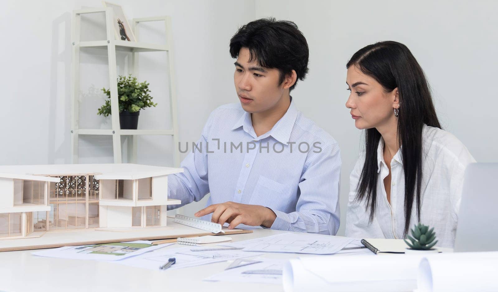 Professional male asian architect using ruler to measure house model length while young beautiful caucasian colleague using laptop to analyzed data on meeting table with house model. Immaculate.