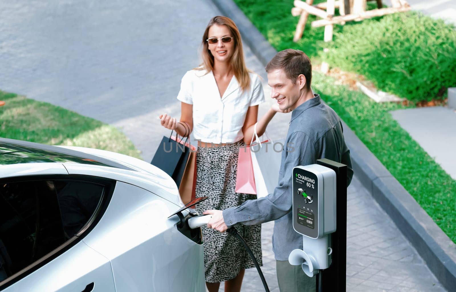 Young couple travel with EV electric car charging in green sustainable city outdoor garden in summer shows urban sustainability lifestyle by green clean rechargeable energy of electric vehicle innards