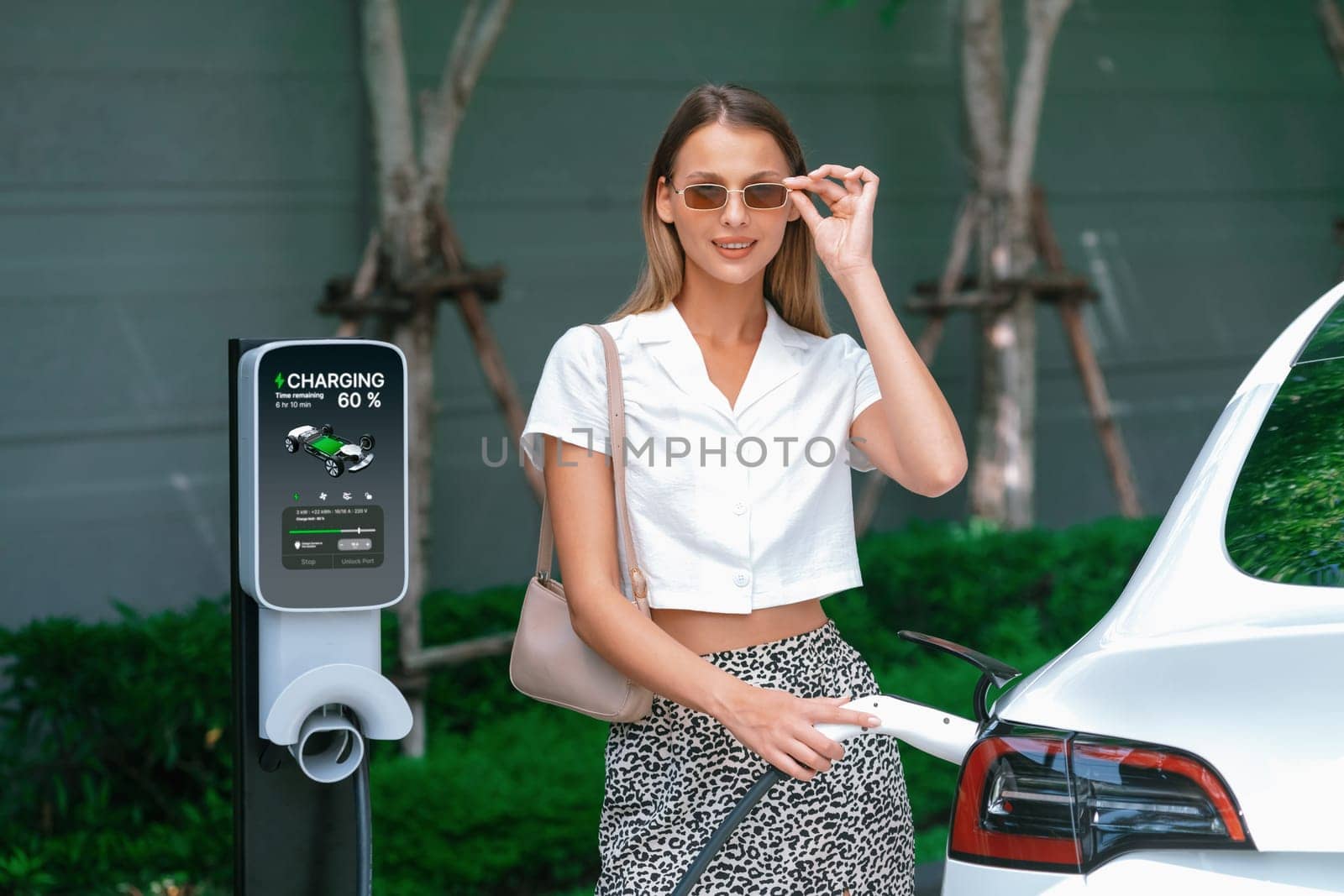 Young woman travel with EV electric car charging in green sustainable city outdoor garden in summer. Urban sustainability lifestyle by green clean rechargeable energy of electric BEV vehicle innards