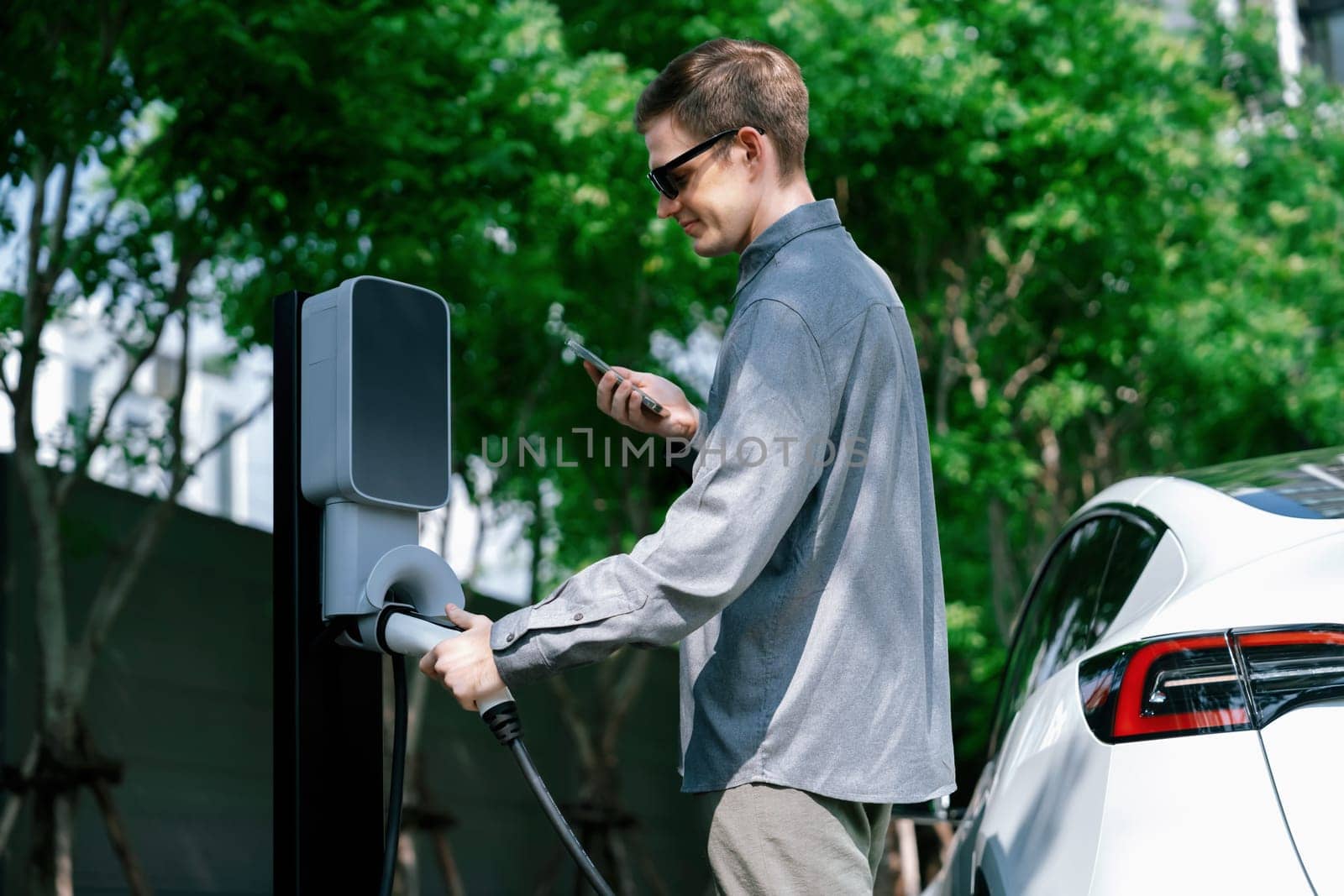 Young man travel with EV electric car charging in green sustainable city outdoor garden in summer shows urban sustainability lifestyle by green clean rechargeable energy of electric vehicle innards