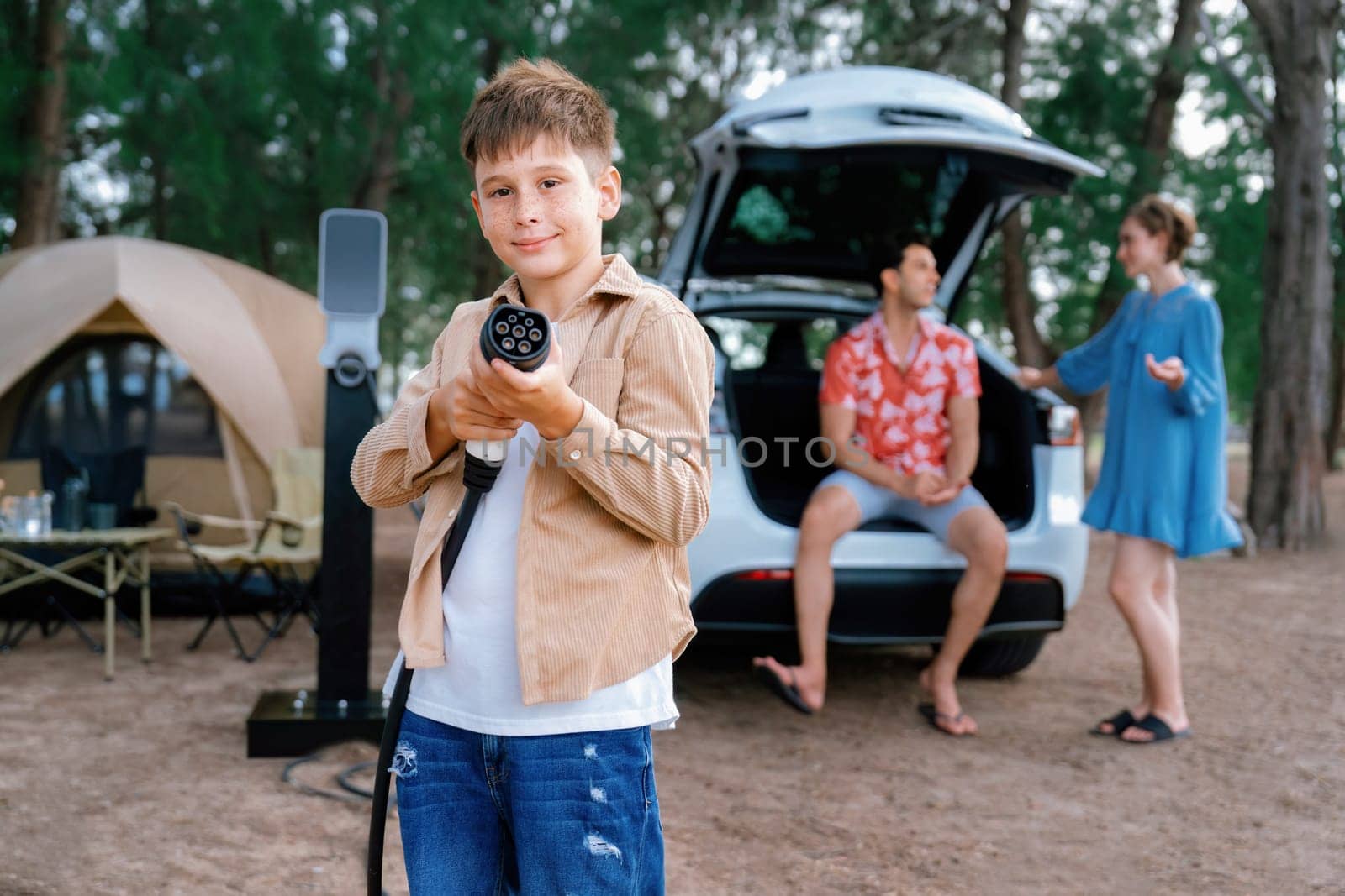 Little boy holding EV charger and point at camera with his family sitting on the trunk in background. Road trip travel with alternative energy charging station for eco-friendly car concept. Perpetual