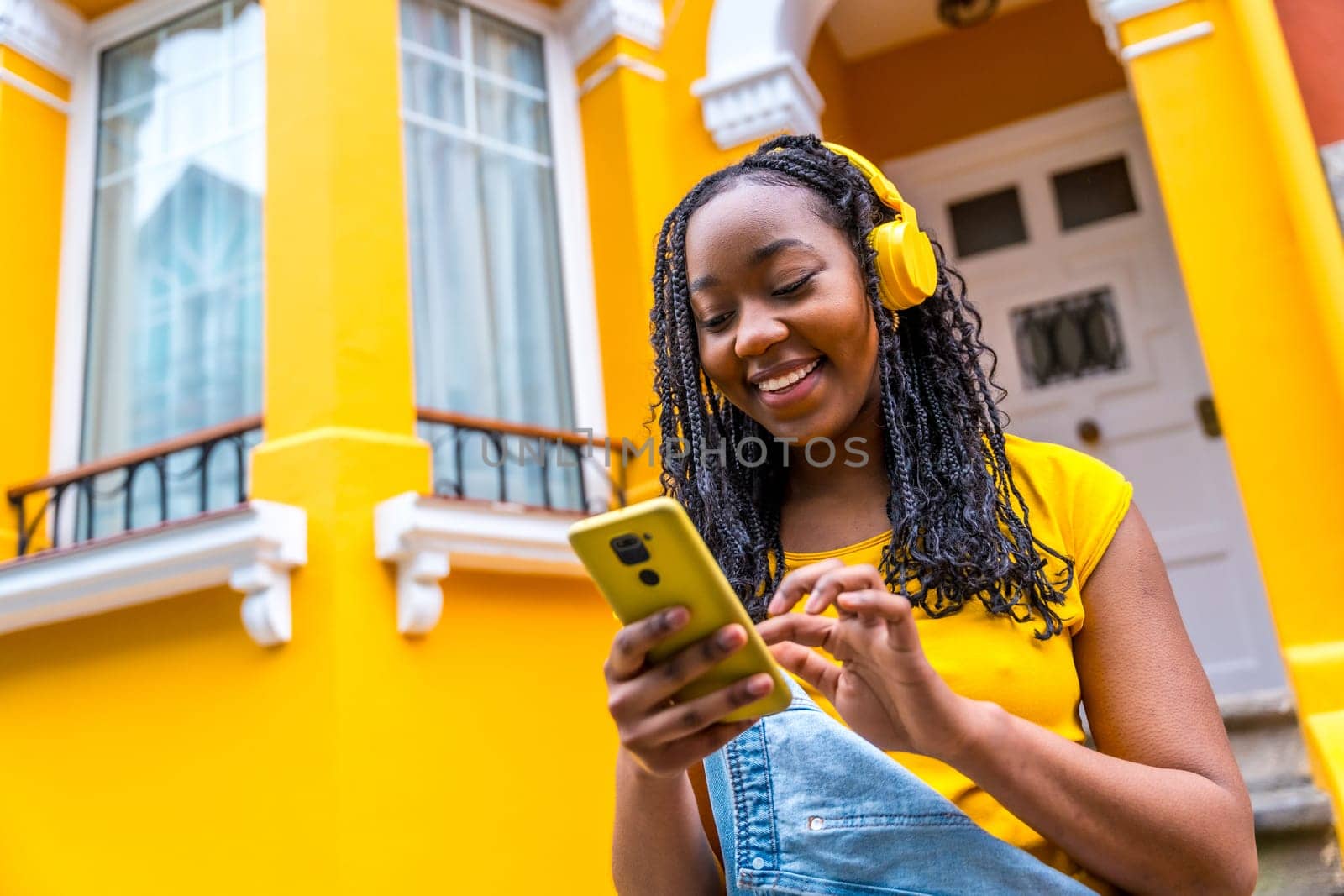 African woman using phone while listening to music standing outside a colorful yellow house