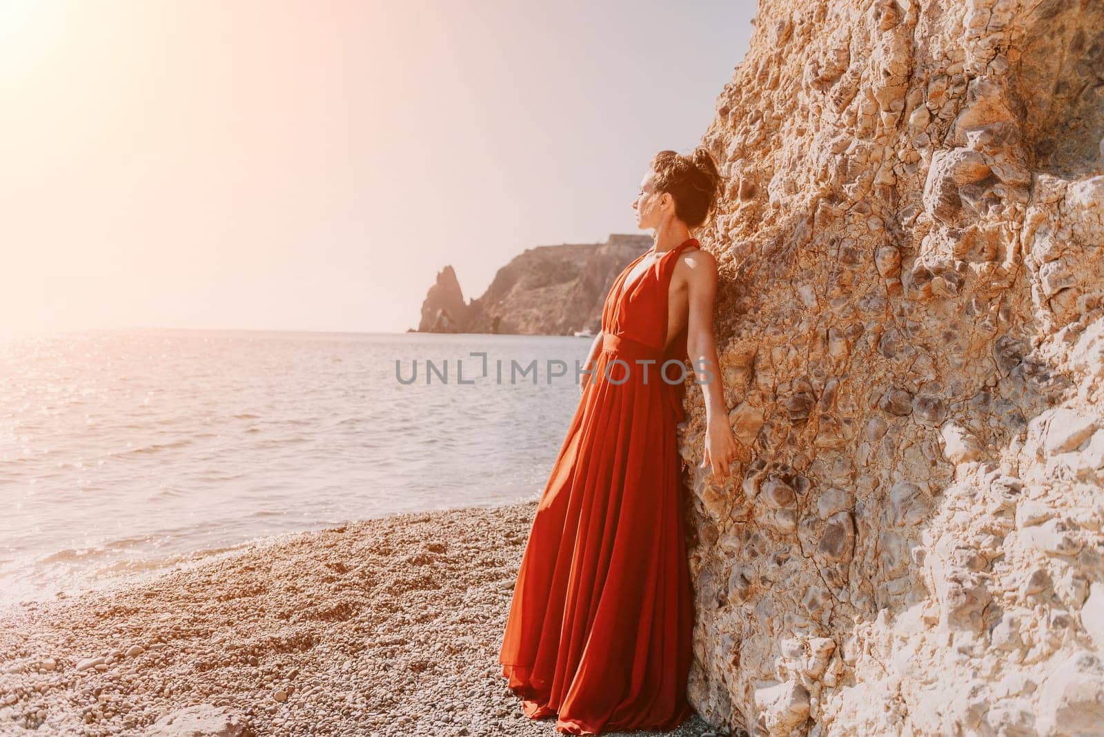 Side view a Young beautiful sensual woman in a red long dress posing on a rock high above the sea during sunrise. Girl on the nature on blue sky background. Fashion photo.