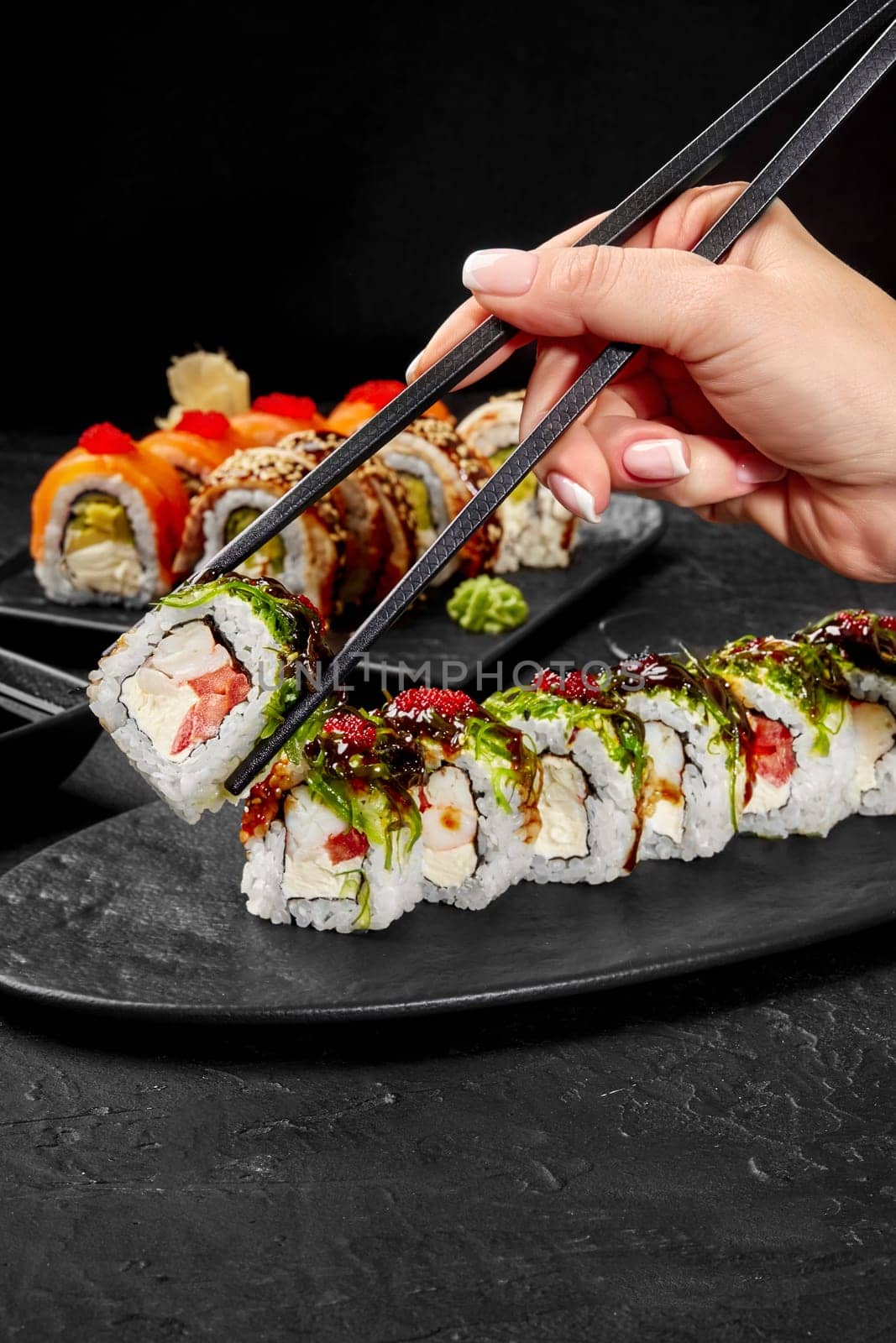 Female hand using traditional Japanese chopsticks to pick up appetizing sushi roll filled with cream cheese, tomato and shrimp topped with wakame seaweed, tobiko and unagi sauce from slate plate