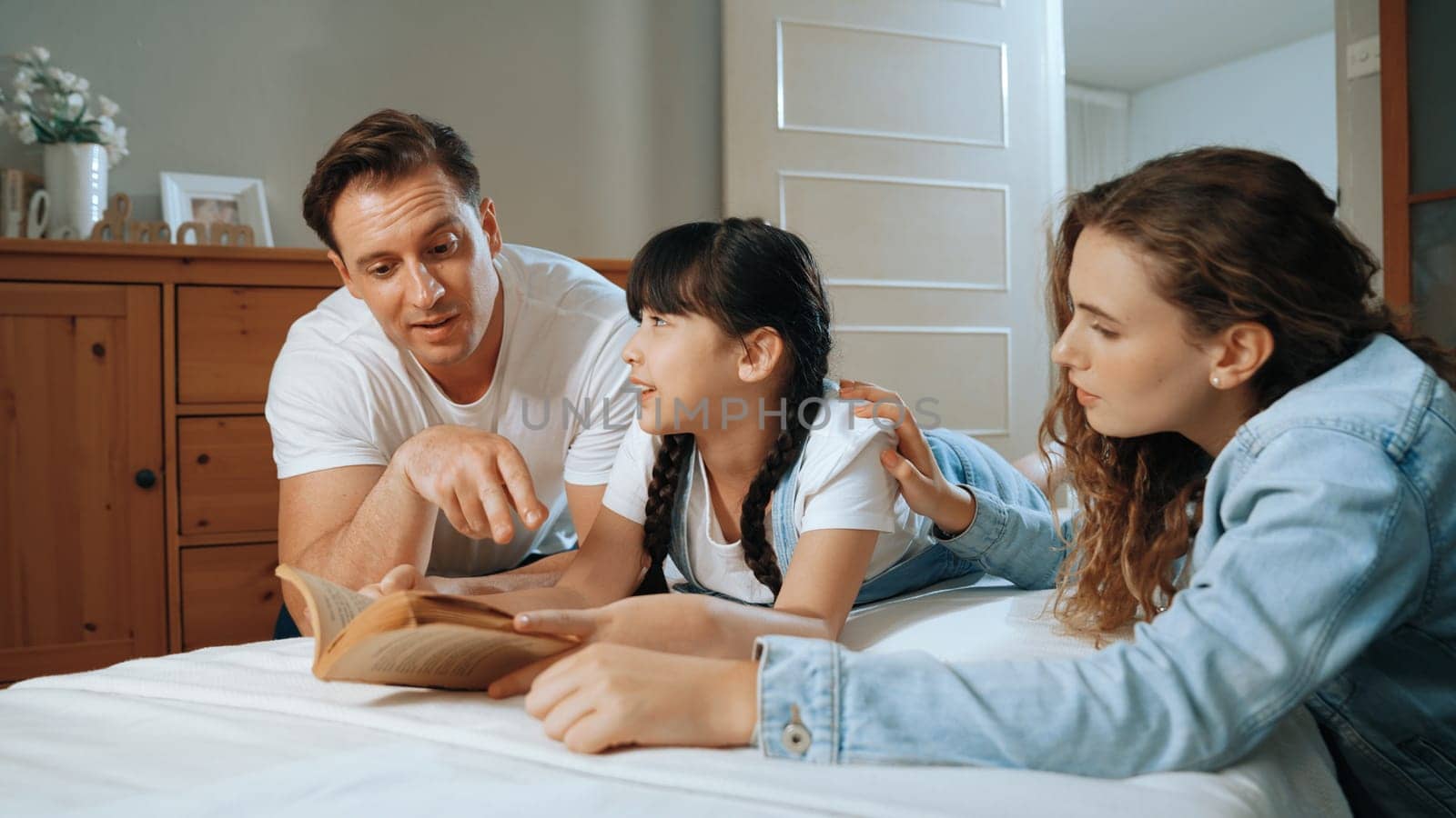 Bedtime story with childhood storytelling, mom and dad reading a fantasy book together to their little young girl in cozy and comfortable bedroom. Modern family happy time. Synchronos