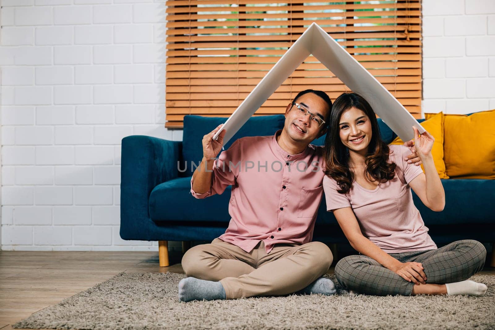 In their new home a smiling Asian couple builds a cardboard house roof signifying happiness and security. Full-length portrait against a white studio wall celebrating achievement and family bonding.