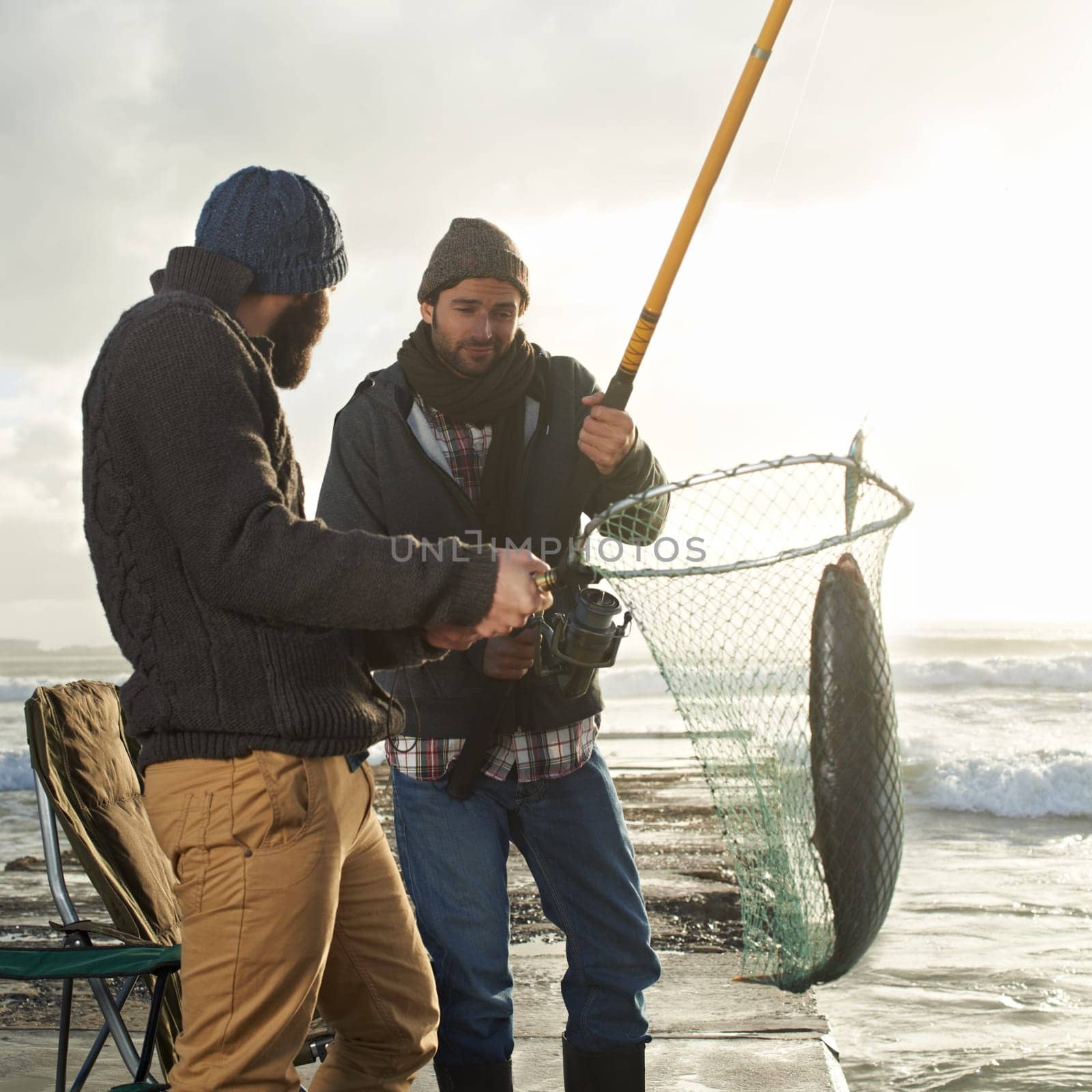 Fishing, men and net with fish at beach with rod, waves and relax on vacation, holiday and adventure. Friendship, people and bonding in morning with overcast, pier and nature for activity and hobby.