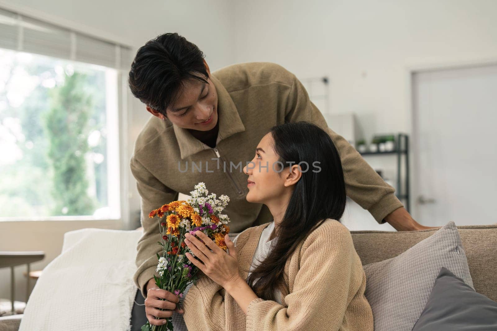 Romantic young asian couple embracing with holding flowers and smiling in living room at home. fall in love. Valentine concept.