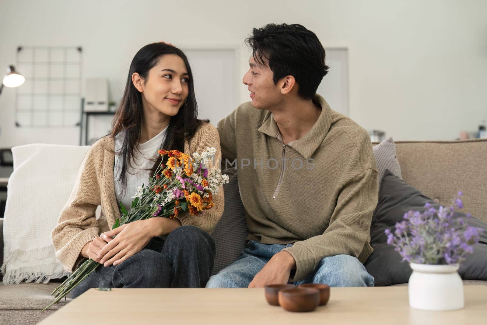 Romantic young asian couple embracing with holding flowers and smiling in living room at home. fall in love. Valentine concept.