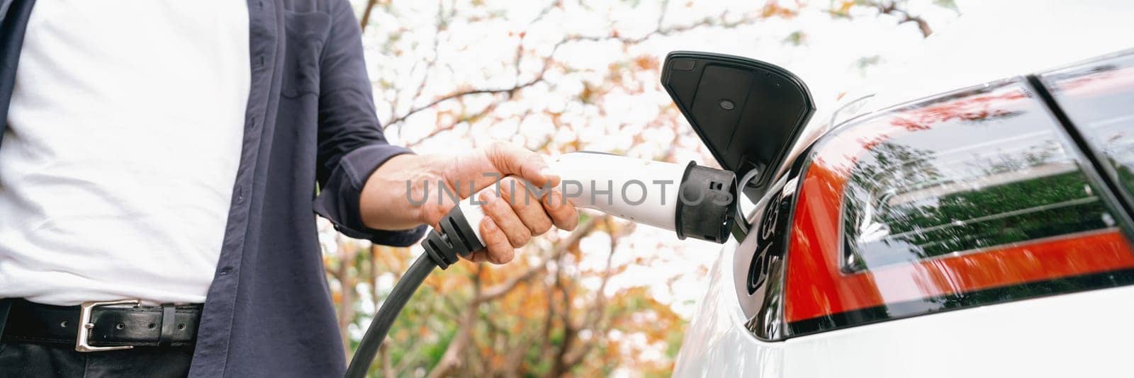 Panorama banner man recharging battery for electric car during autumnal road trip travel EV car in autumnal forest. Eco friendly travel on vacation during autumn with electric vehicle. Exalt