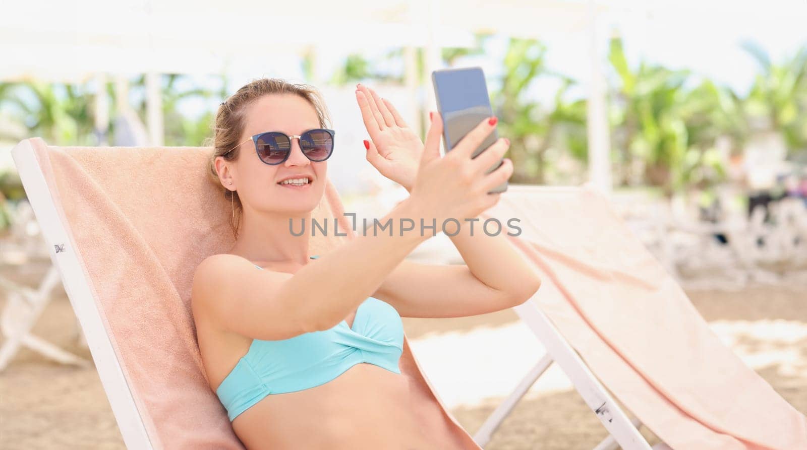 A woman in a swimsuit lies on a sun lounger, takes a selfie, close-up. Phone call via video link, photo from vacation