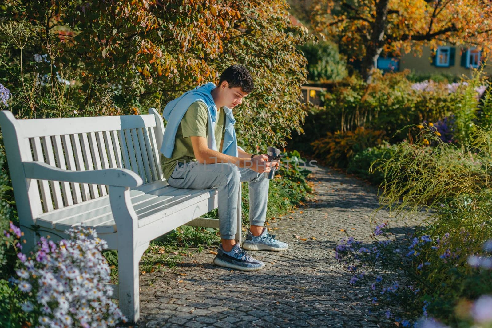 A Teenager Sits On A Bench In The Autumn Park Drinks Coffee From A Thermo Mug And Looks Into A Phone. Portrait Of Handsome Cheerful Guy Sitting On Bench Fresh Air Using Device Browsing Media Smm Drinking Latte Urban Outside Outdoor by Andrii_Ko