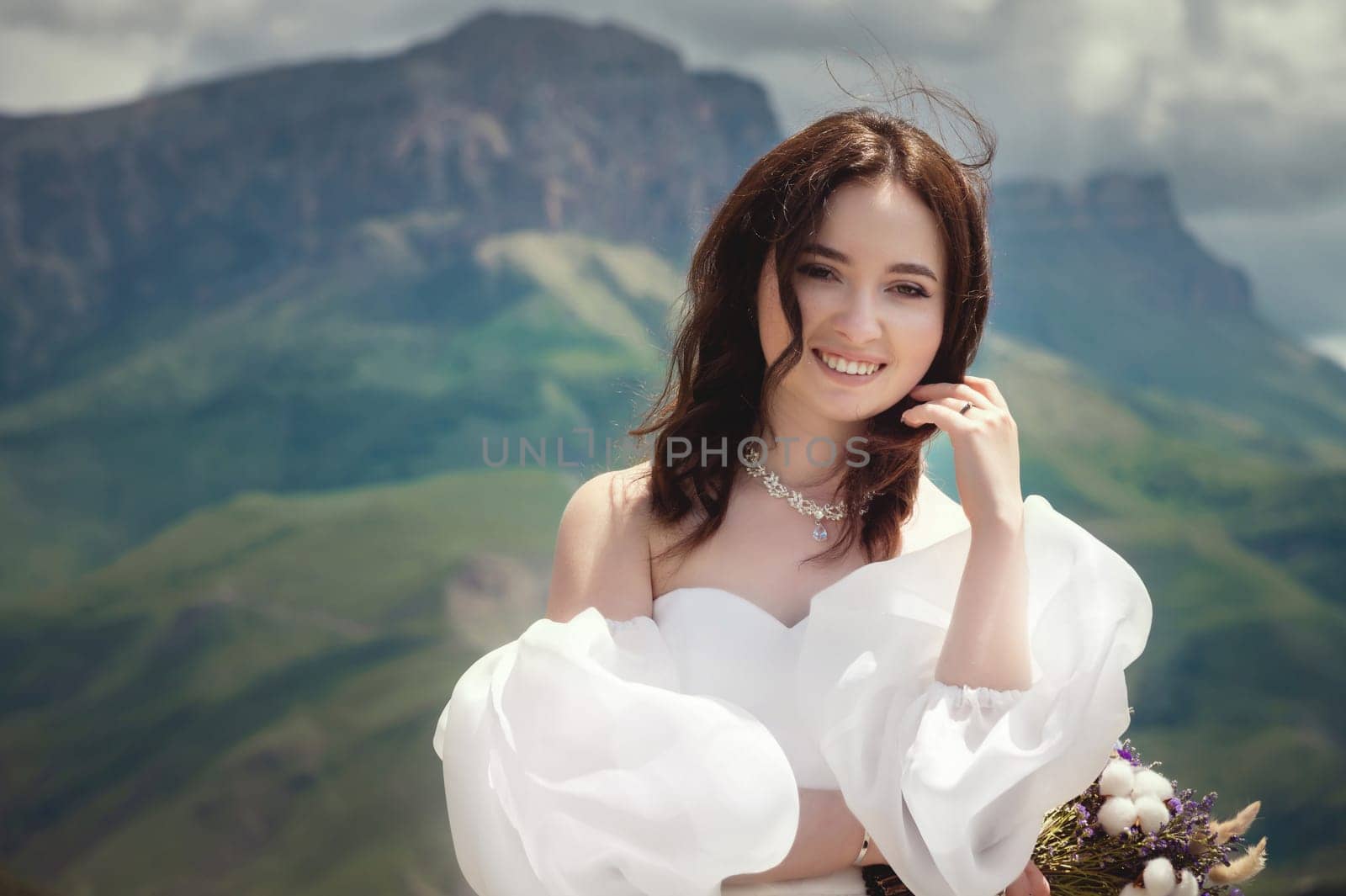 The bride with a wedding bouquet, her eyes closed and her face basking in the sun in the mountains. panoramic view of mountain range from behind of married young woman.