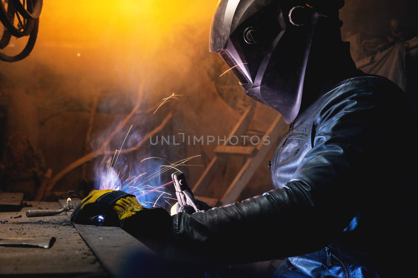 A male welder in a leather jacket carries out welding work in a home shed against the background of a brick wall. Handicraft production of metal structures and products.