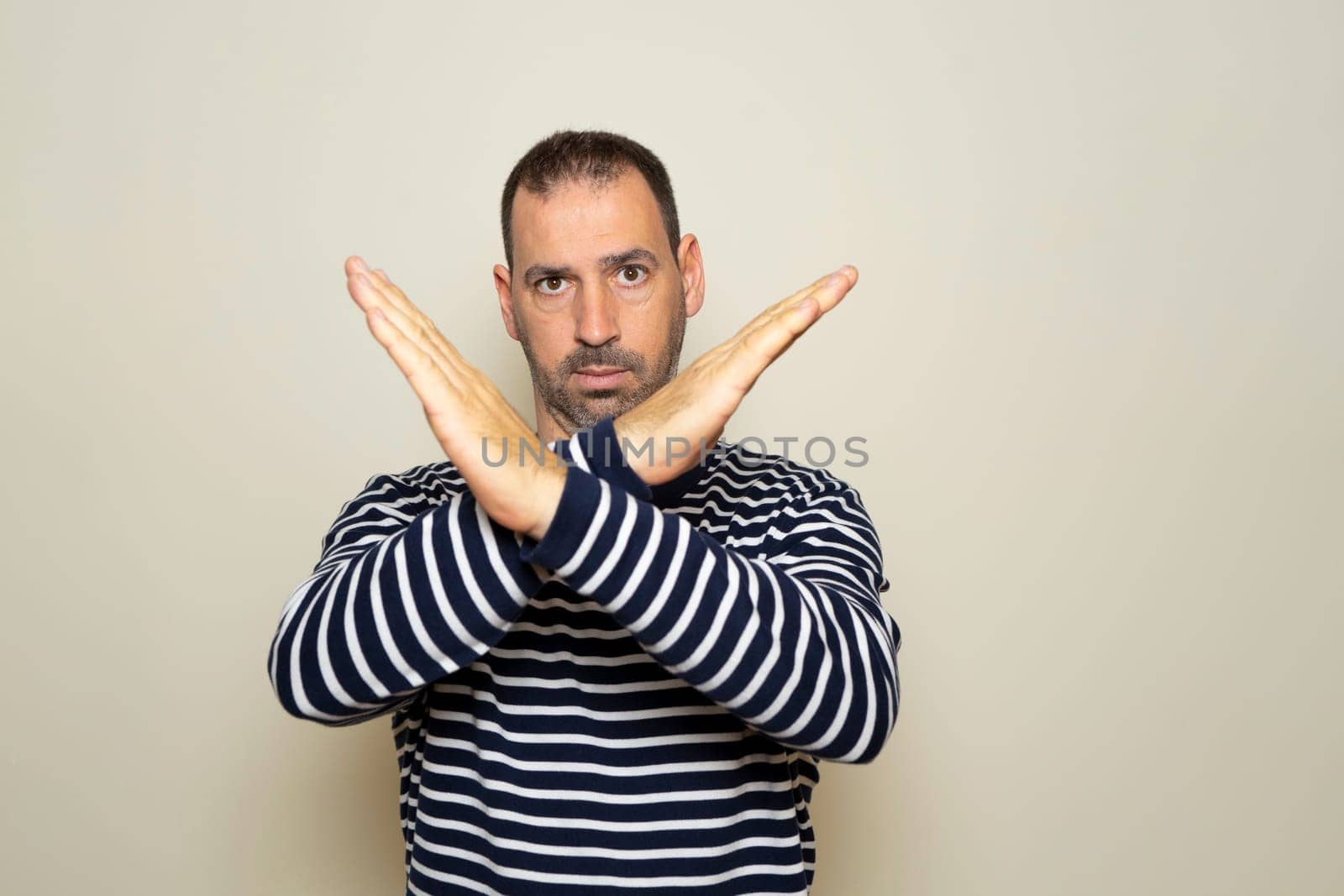 Portrait of serious Hispanic man with hands crossed, showing x sign which means stop, warning of trouble. Isolated on beige studio background. by Barriolo82