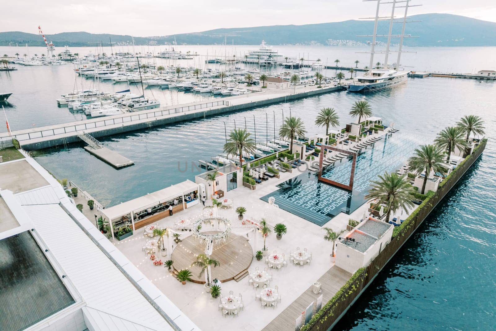 Round festive tables stand around a stage by the long pool on the pier of a luxurious marina. High quality photo