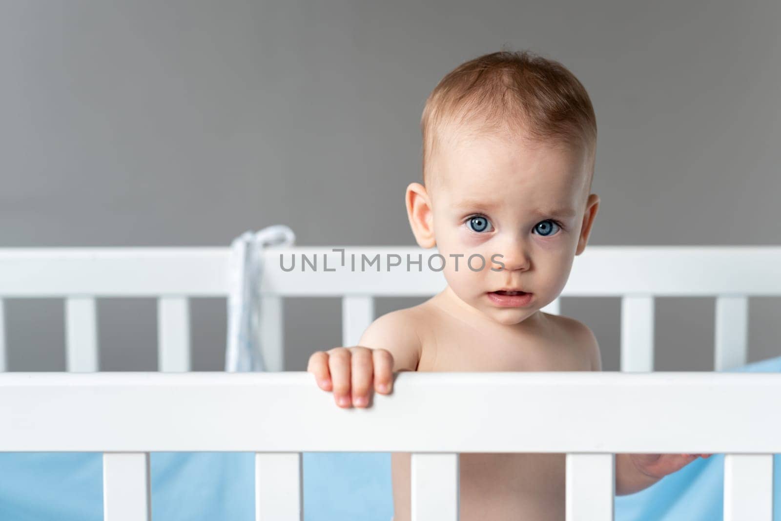 Sad baby girl stands gripping the rail of a white wooden crib, her eyes reflecting a sense of