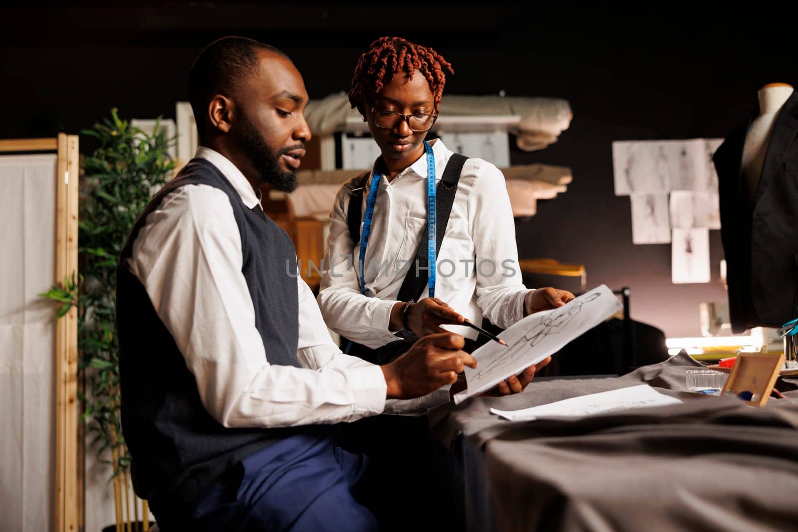 Brainstorming apprentice tailors looking over sketch drawings of bespoke collection imagined by master fashion designer. Teamworking atelier shop colleagues discussing sartorial adjustments to be made