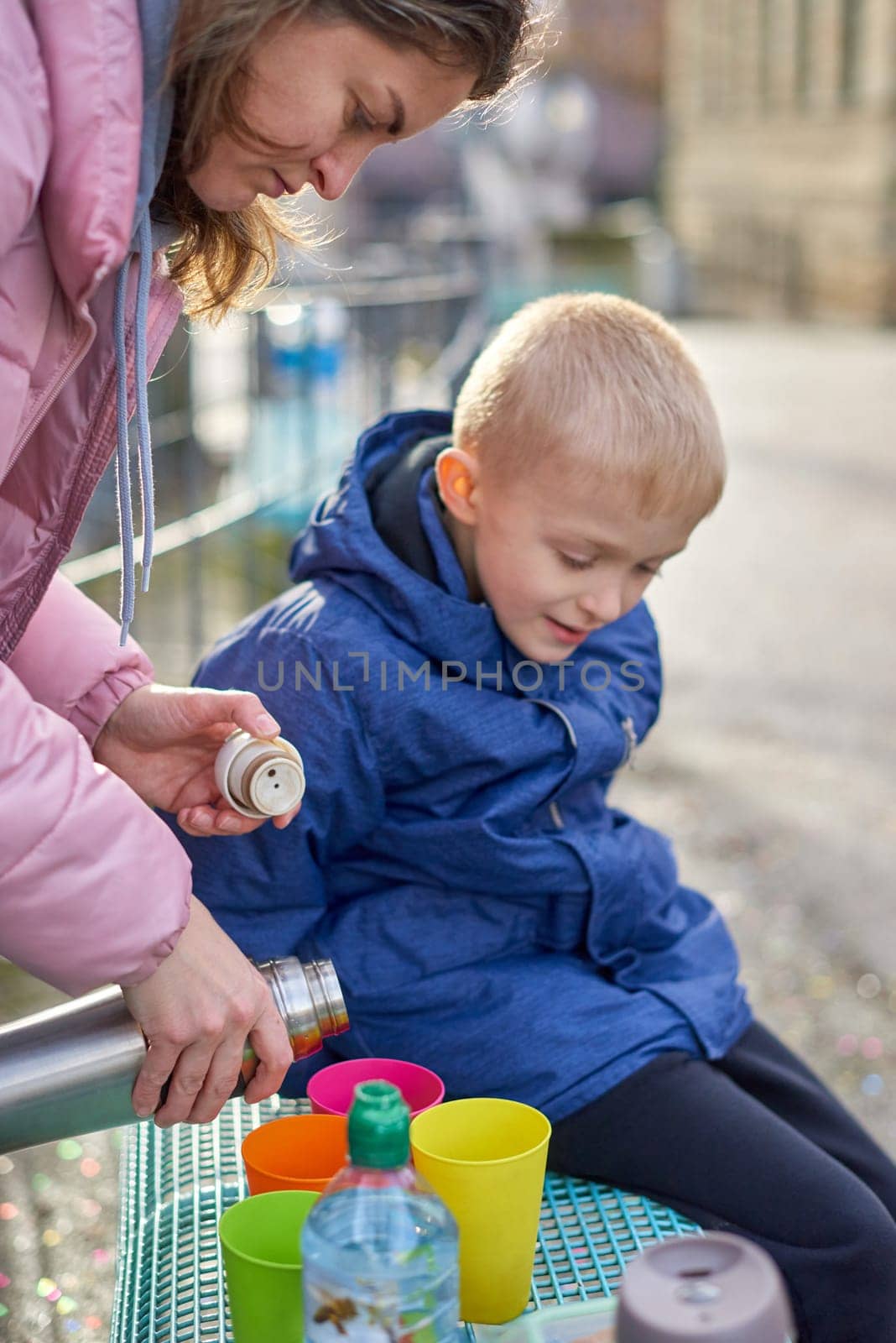 Family Picnic Delight: Cheerful 8-Year-Old Blond Boy in Blue Winter Jacket Sits on Bench While Mom Pours Tea from Thermos, Autumn or Winter. a joyful 8-year-old blond boy in a blue winter jacket, sitting on a bench while his mom pours tea from a thermos into colorful plastic cups. by Andrii_Ko