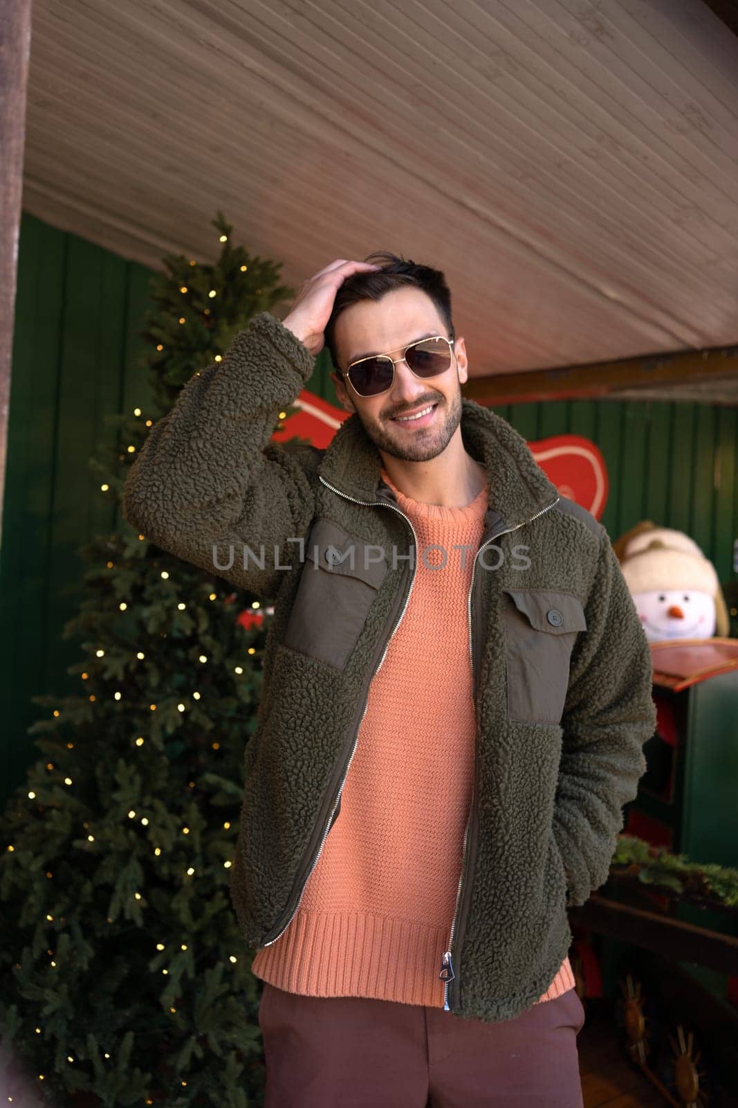 Portrait of happy handsome man in trendy jacket and sunglasses standing near Christmas tree. Fashionable male hipster posing with hand in hair and smiling.