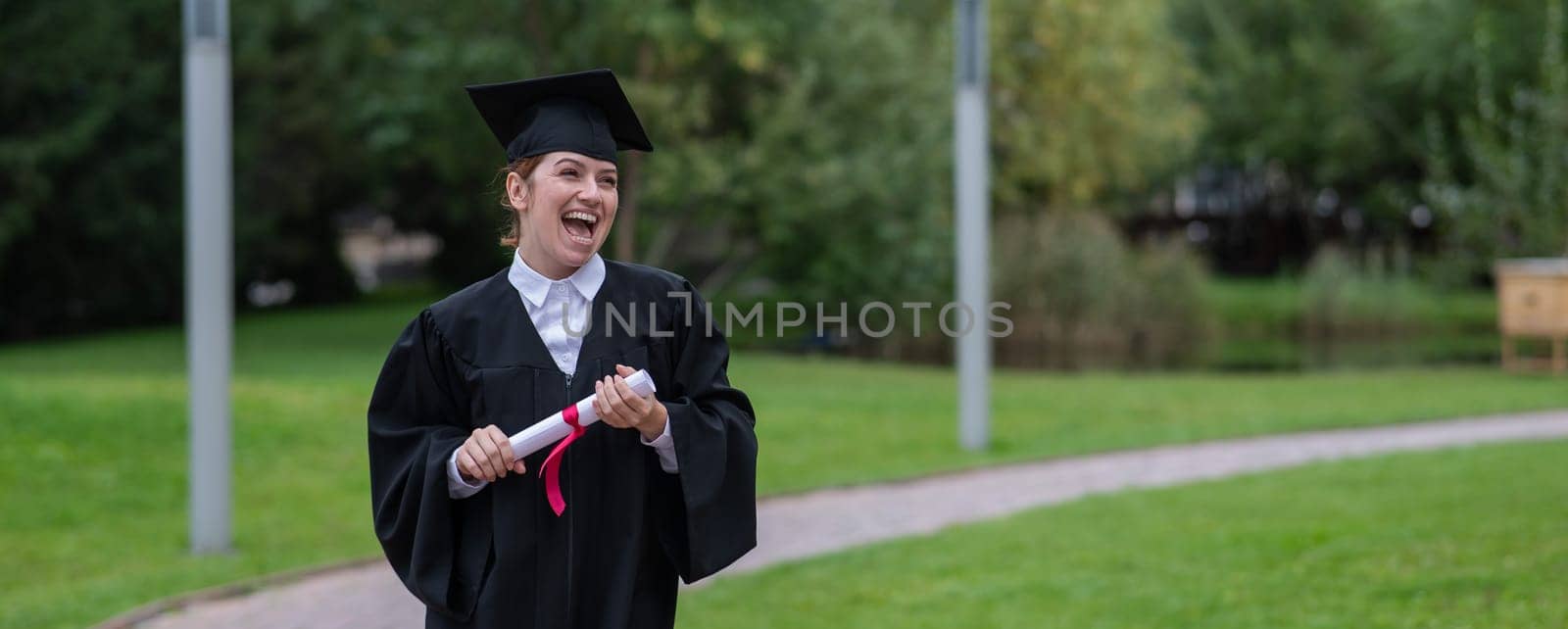 Portrait of happy caucasian woman in graduate gown holding diploma outdoors. by mrwed54