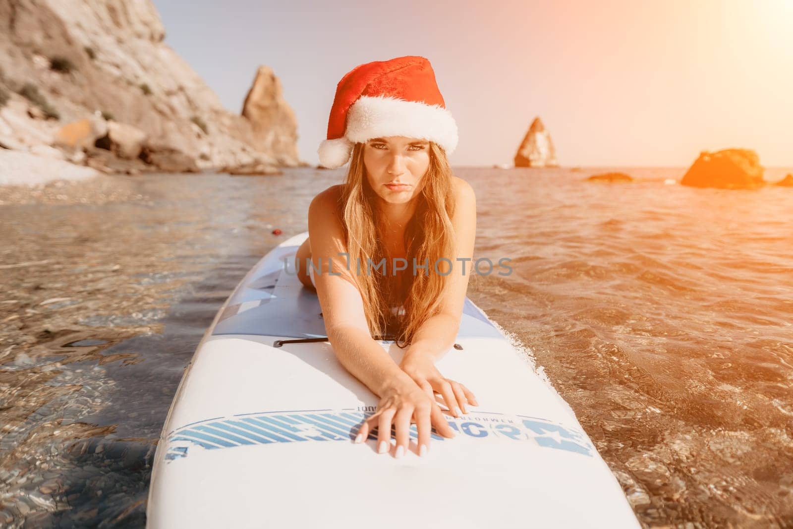 Woman sea sup. Close up portrait of happy young caucasian woman with long hair in Santa hat looking at camera and smiling. Cute woman portrait in a white bikini posing on sup board in the sea by panophotograph