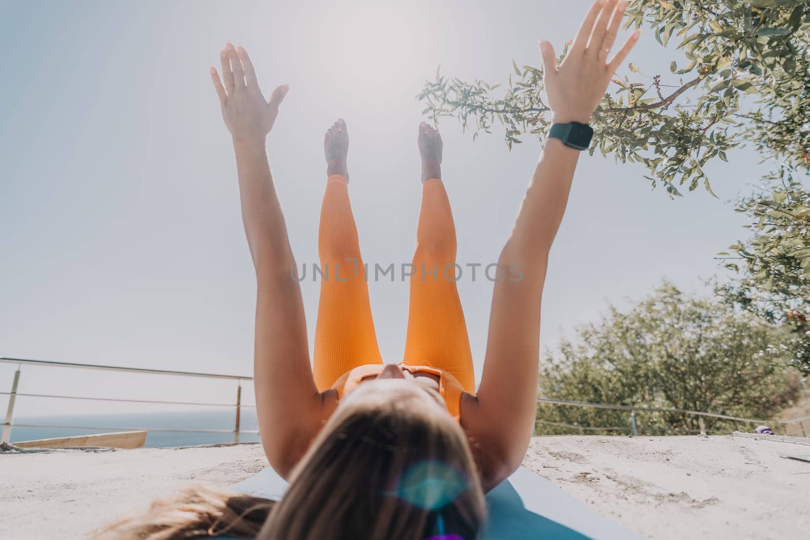 Fitness woman sea. Happy middle aged woman in orange sportswear exercises morning outdoors on yoga mat with laptop in park over ocean beach. Female fitness pilates yoga routine. Healthy lifestyle. by panophotograph