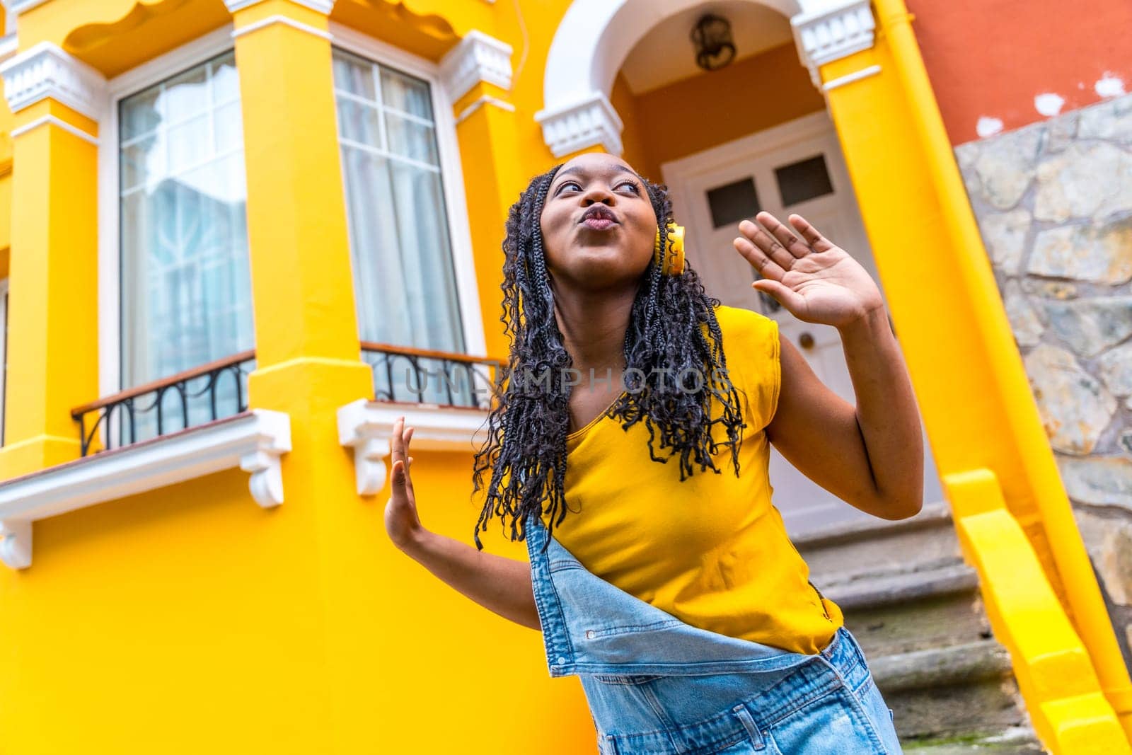 Cute african woman dancing listening to music with headphones next to a yellow house outdoors