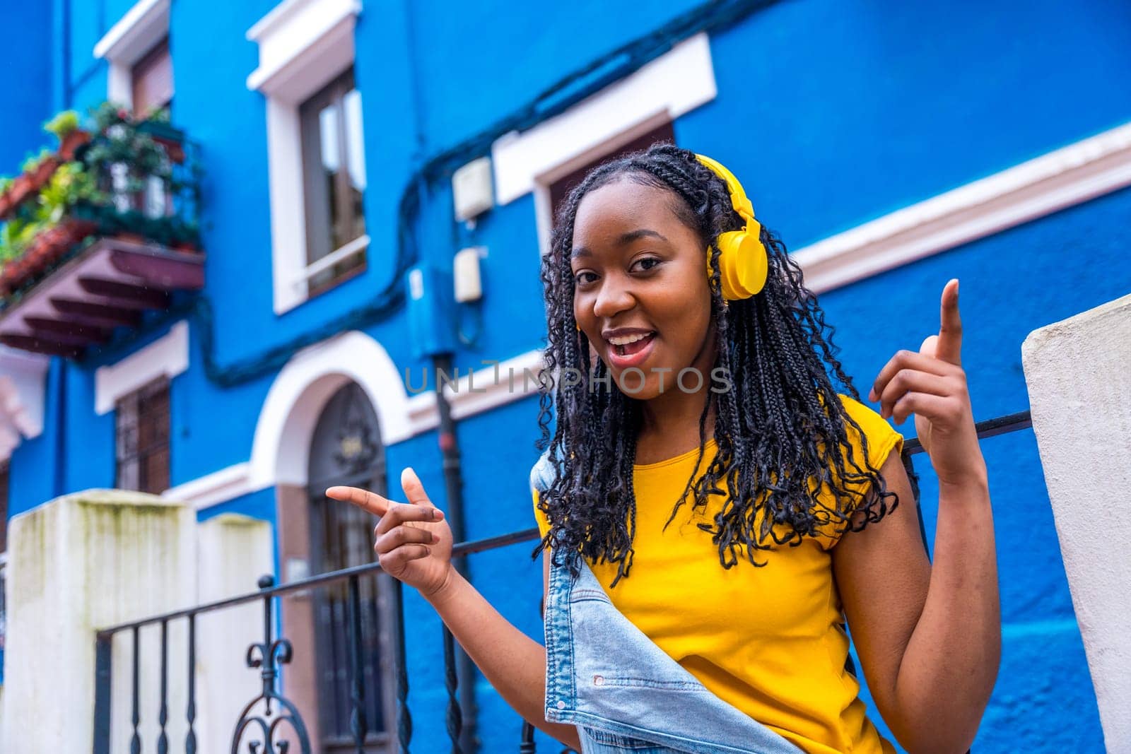 African woman singing while listening to music with headphones standing in a street with blue colored houses