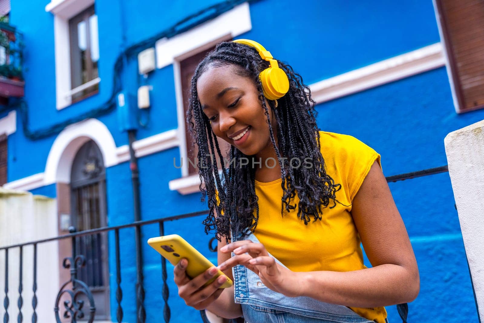 Smiling african woman using phone and listening to music with headphones outside a blue house