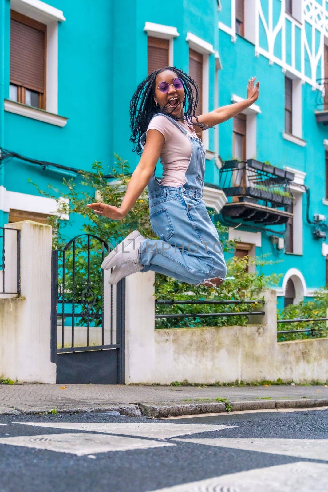 African young woman jumping in a street with colorful houses by Huizi