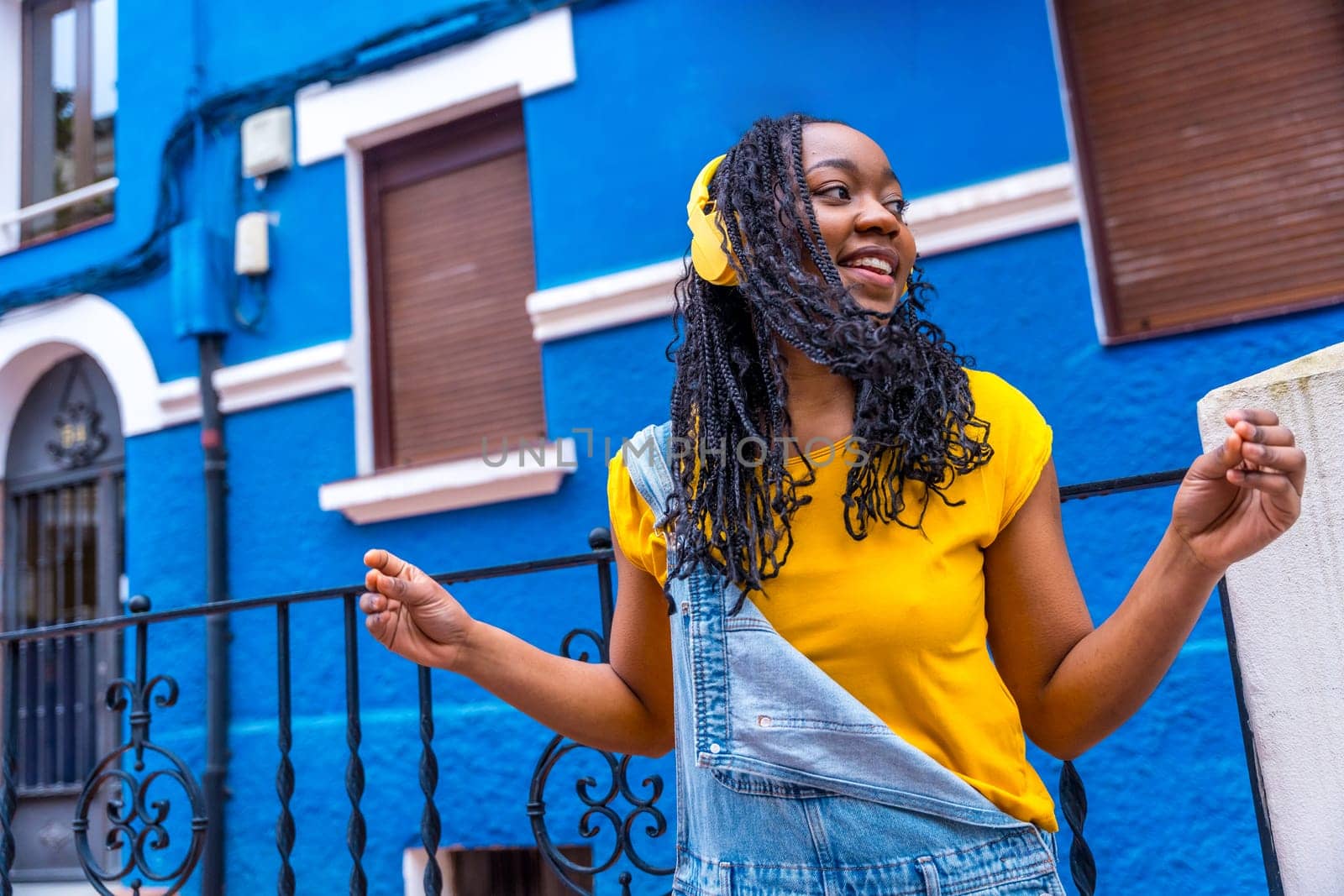 Cool casual african woman with braided hair dancing listening to music with headphones outdoors next to a blue house