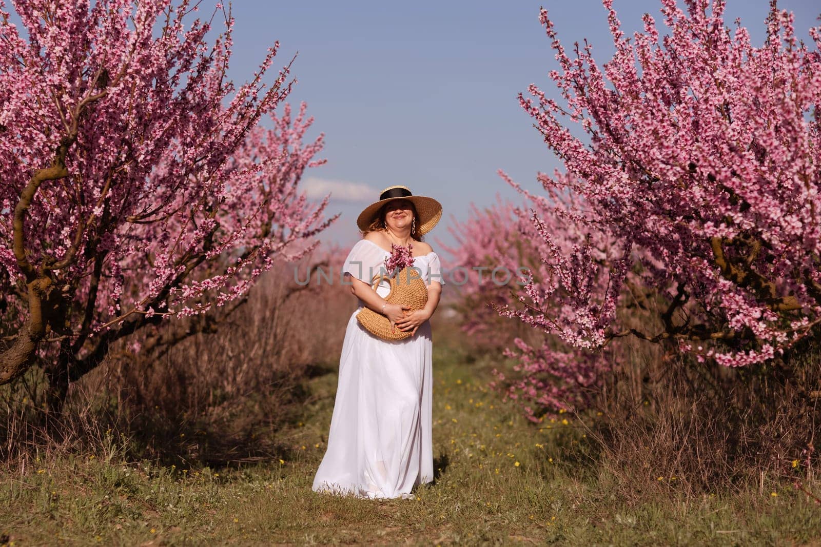 Woman blooming peach orchard. Against the backdrop of a picturesque peach orchard, a woman in a long white dress and hat enjoys a peaceful walk in the park, surrounded by the beauty of nature. by Matiunina