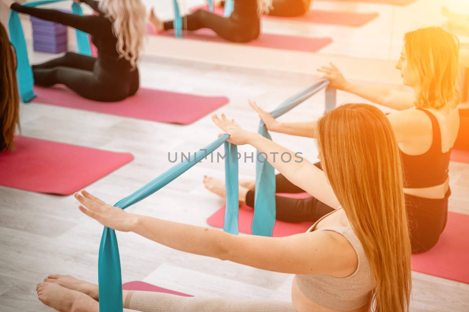 A group of six athletic women doing pilates or yoga on pink mats in front of a window in a beige loft studio interior. Teamwork, good mood and healthy lifestyle concept. by Matiunina