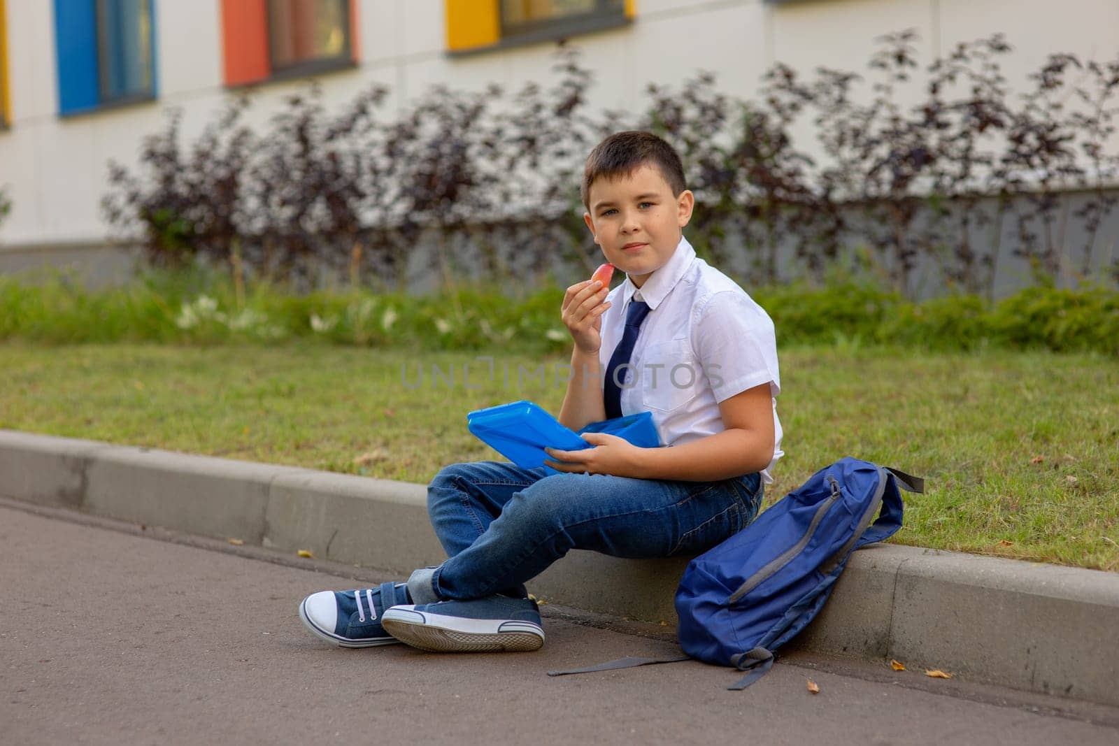 schoolboy in a white shirt with a blue tie, holds a blue lunch box and a slice of apple, looks at the camera.