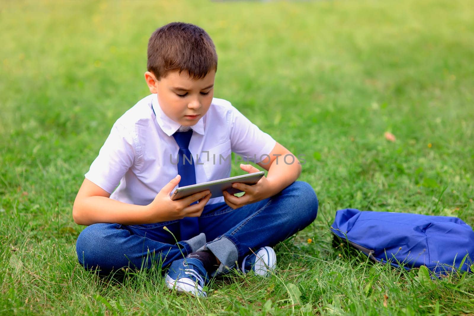 Serious Schoolboy in a white shirt and blue tie sits on green grass against a blue sky with white clouds and looks into a tablet. Copy space. Close up