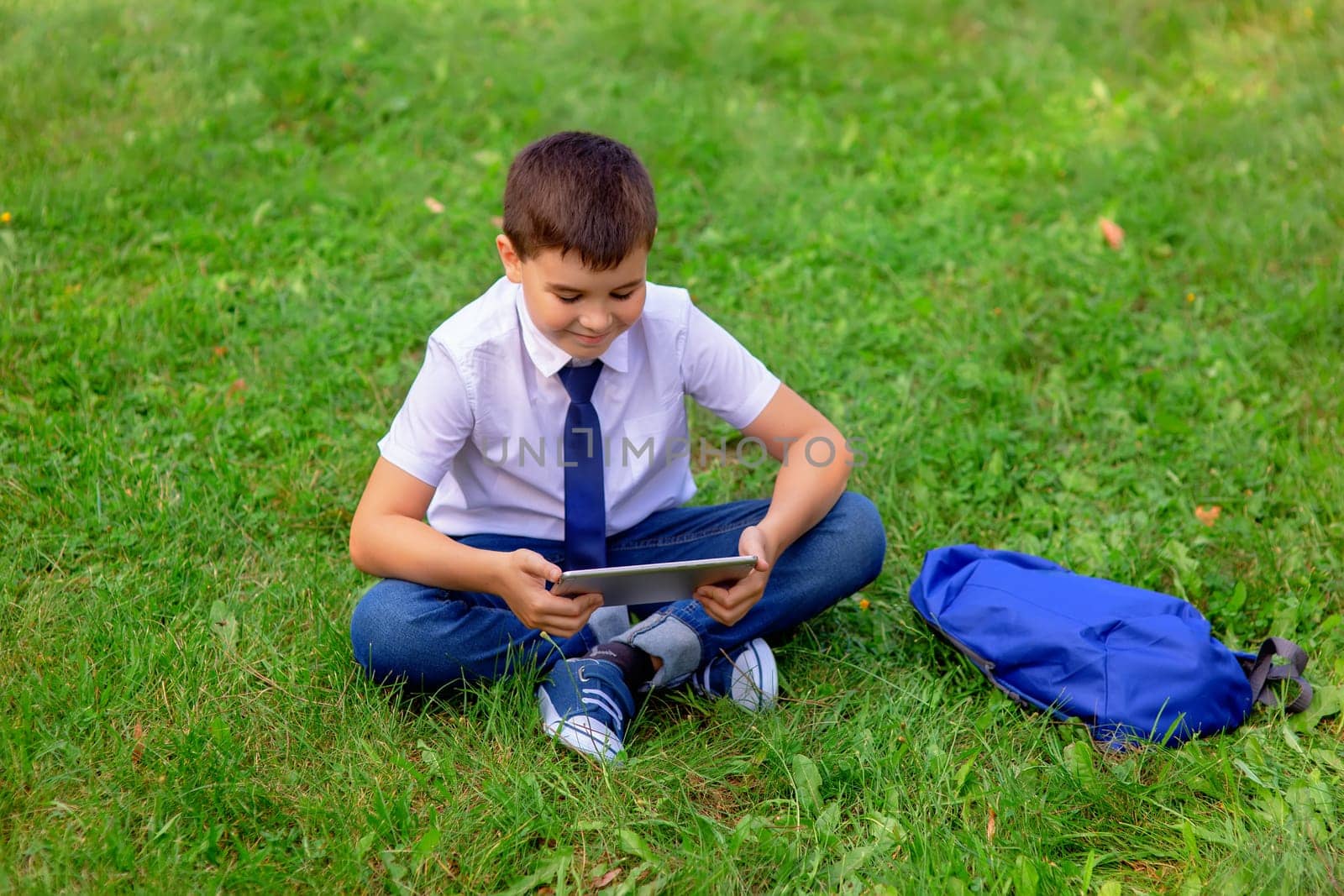 Happy Schoolboy in a white shirt and blue tie sits on green grass against a blue sky with white clouds and looks into a tablet. Copy space. Top view