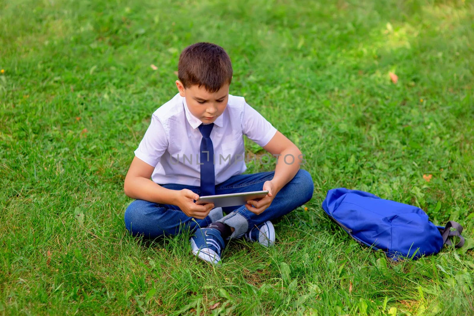 Serious Schoolboy in a white shirt and blue tie sits on green grass against a blue sky with white clouds and looks into a tablet. Copy space. Top view