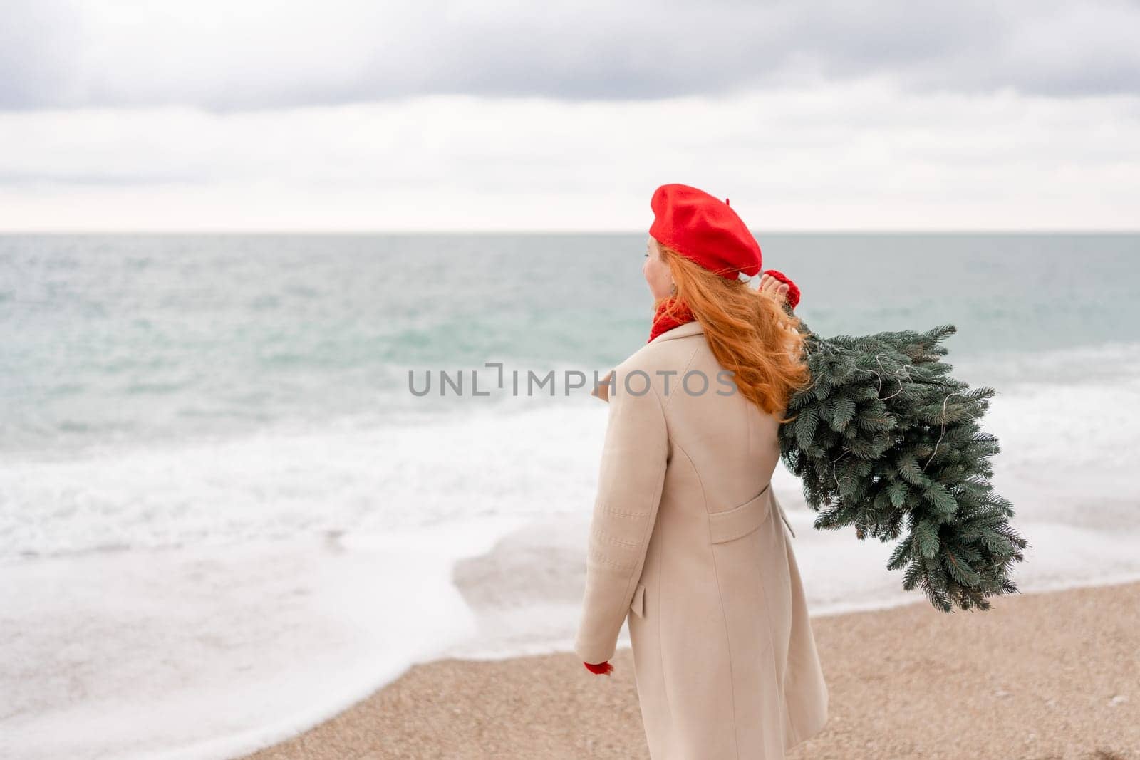 Redhead woman Christmas tree sea. Christmas portrait of a happy redhead woman walking along the beach and holding a Christmas tree on her shoulder. She is dressed in a light coat and a red beret