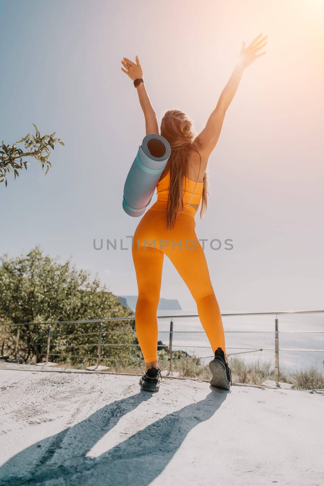 Fitness woman sea. Happy middle aged woman in orange sportswear exercises morning outdoors on yoga mat with laptop in park over ocean beach. Female fitness pilates yoga routine. Healthy lifestyle. by panophotograph