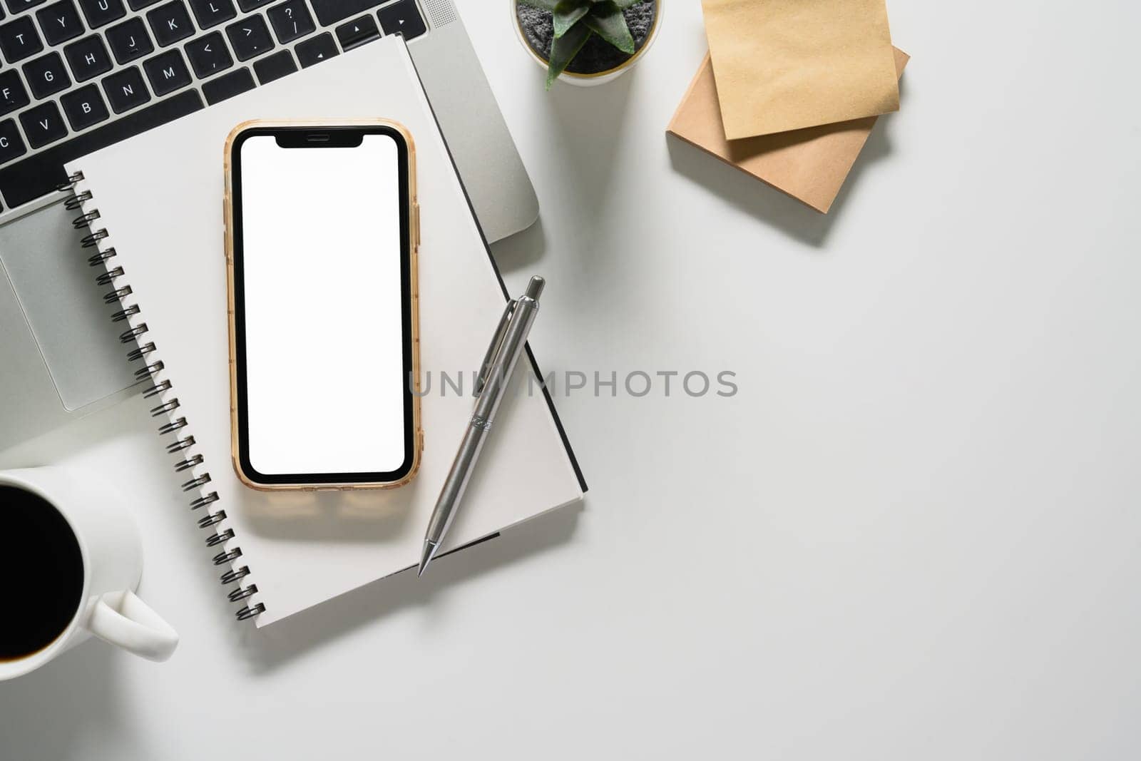 Smartphone with blank screen, notepad, coffee cup and laptop on white table by prathanchorruangsak