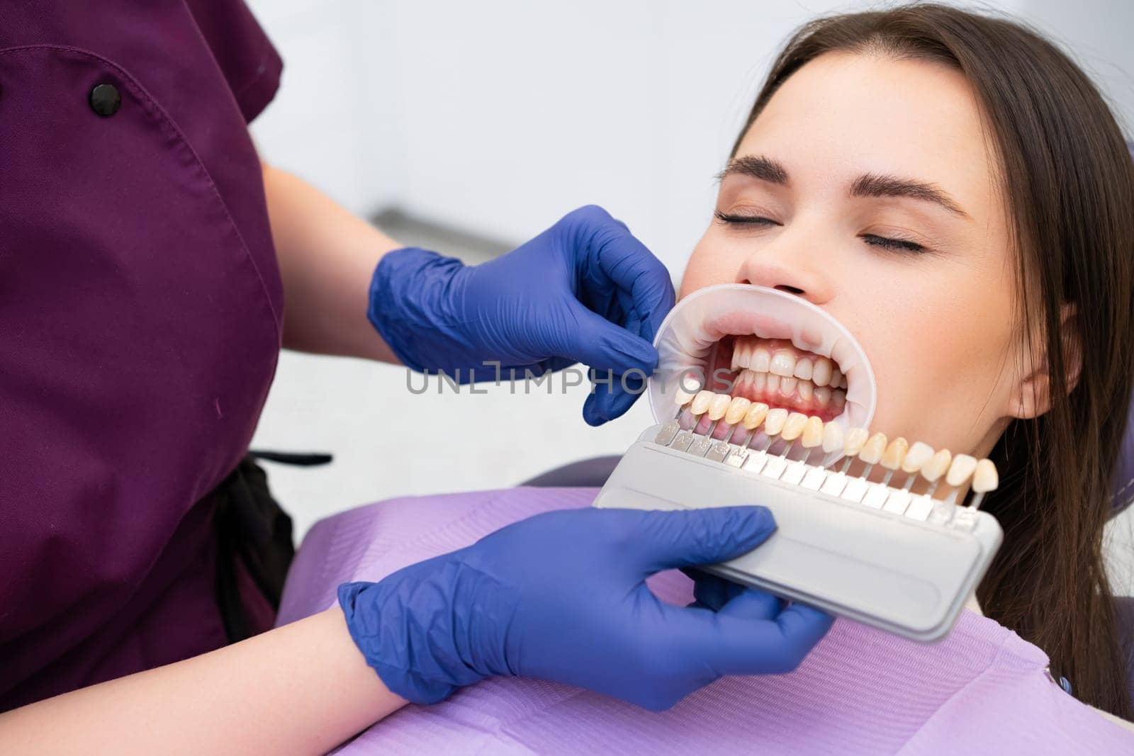 The dentist carefully selects the enamel shade for the young brunette womans teeth before beginning the whitening procedure.