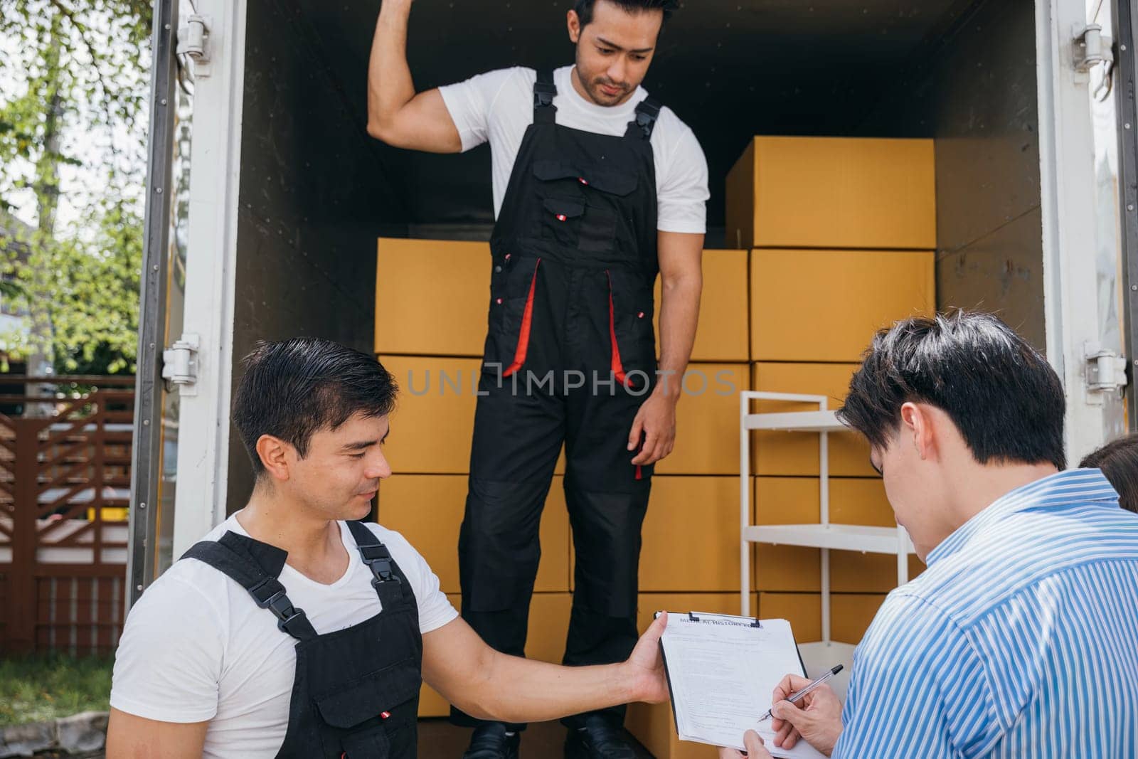 Satisfied couple signs delivery checklist after professional movers aid in furniture lifting. Uniformed employees emphasize teamwork for customer contentment. Moving Day Concept by Sorapop
