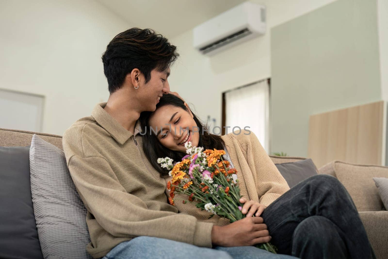 Romantic young asian couple embracing with holding flowers and smiling in living room at home. fall in love. Valentine concept.