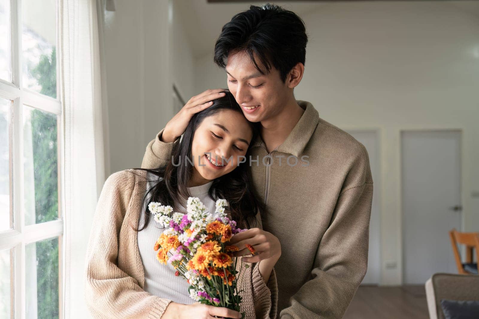 Romantic young asian couple embracing with holding flowers and smiling in living room at home. fall in love. Valentine concept.