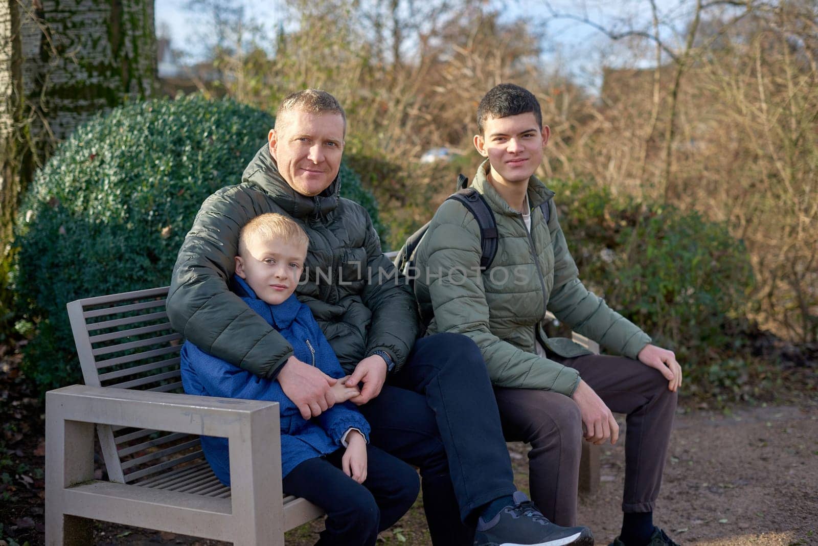 Dad with two sons sitting on a bench in autumn park. Experience the tranquility of familial bonds in the heart of autumn with this serene image. A father, 40 years old, and his two sons - a beautiful 8-year-old boy and a 17-year-old young man, seated in the park. The autumnal ambiance adds warmth to this captivating moment of family togetherness.