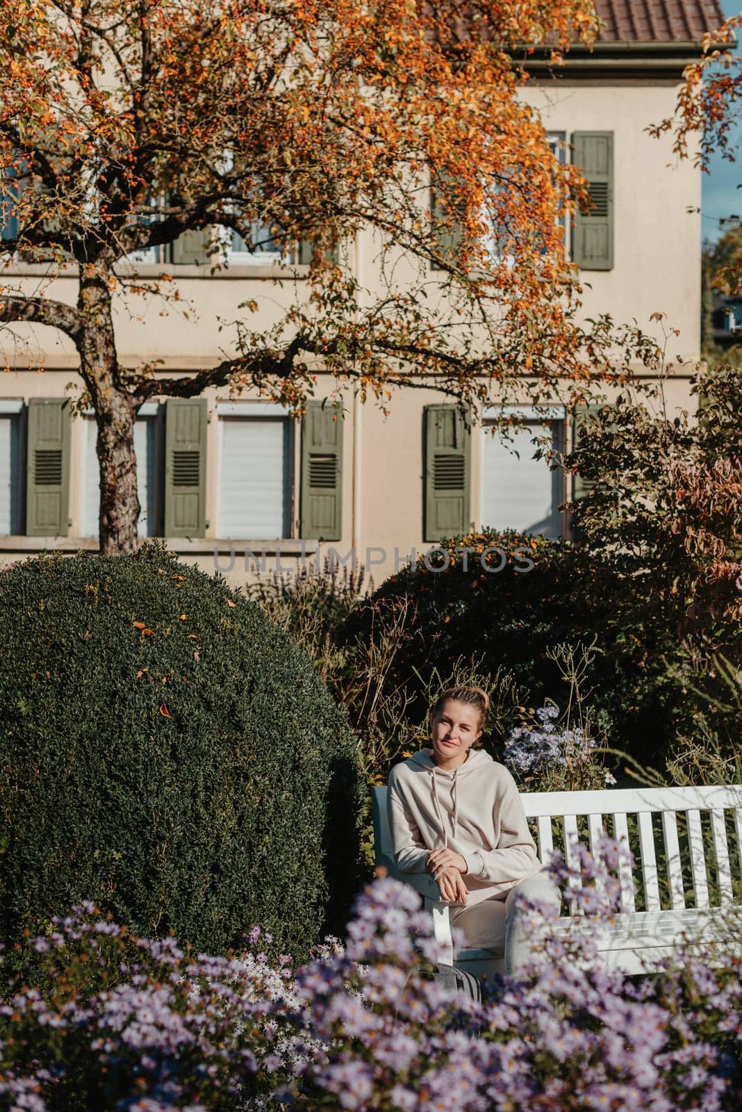 A Girl Sits On A Bench In The Park And Enjoys The Sun. Portrait Young Adult Attractive Woman Enjoy Sitting On Bench And Relaxing Calm Carefree Rest In City Park Against Green Grass And Trees On Sunny Day. Single Female Person Relaxing Chilling Outdoors by Andrii_Ko