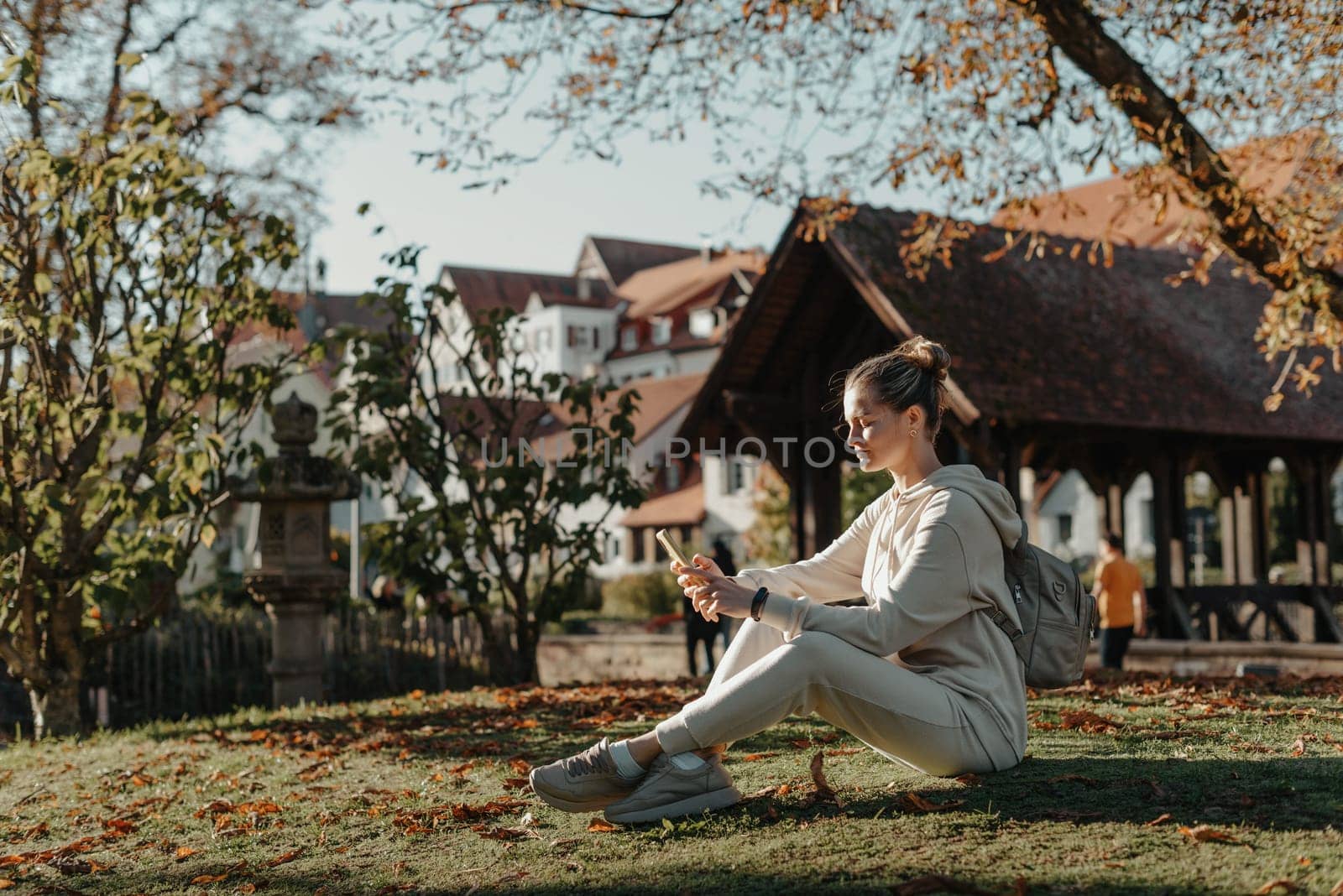 Young Fashionable Teenage Girl With Smartphone In Europian Park In Autumn Sitting At Smiling. Trendy Young Woman In Fall In Park Texting. Retouched, Vibrant Colors. Beautiful Blonde Teenage Girl Wearing Casual Modern Autumn Outfit Sitting In Park In Autumn. Retouched, Vibrant Colors, Brownish Tones. by Andrii_Ko